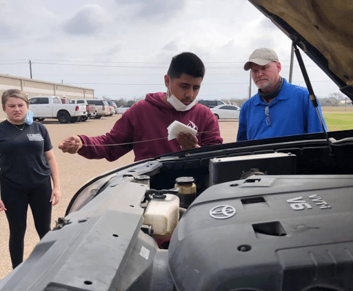 Hybrid school student learning to check oil on a car.